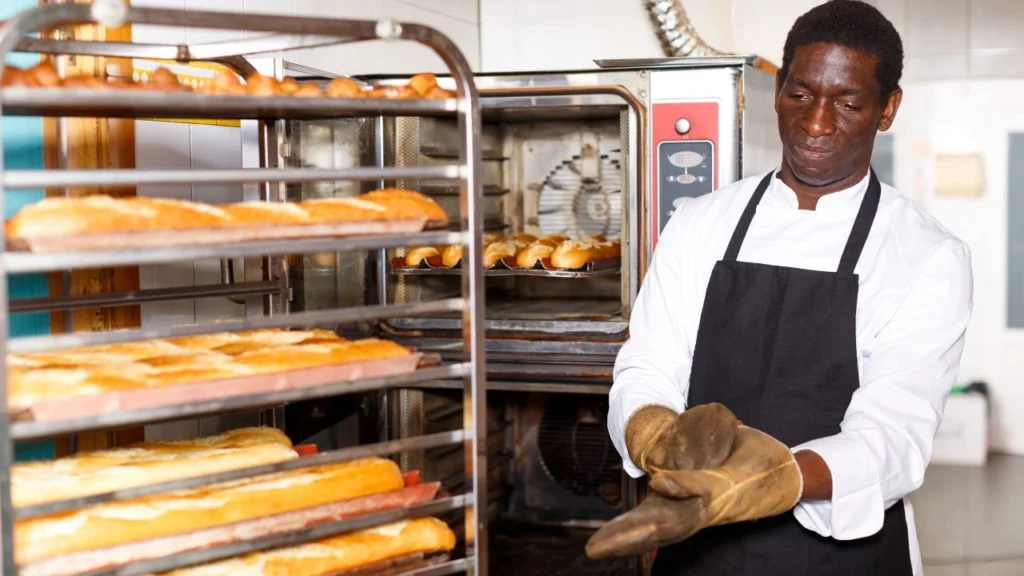Baker wearing heat-resistant gloves near bread racks