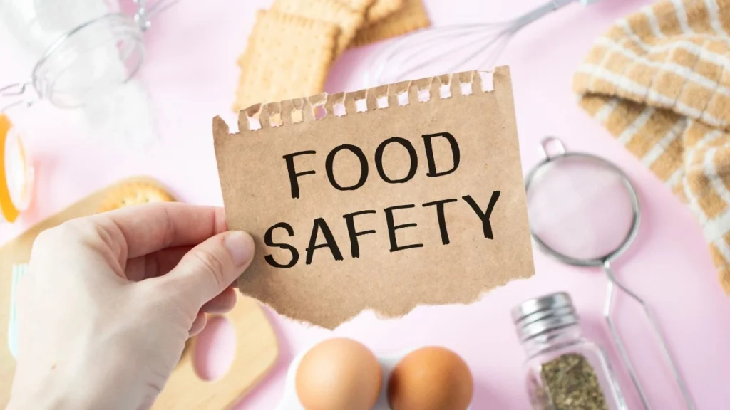 Hand holding 'Food Safety' sign with kitchen tools background.