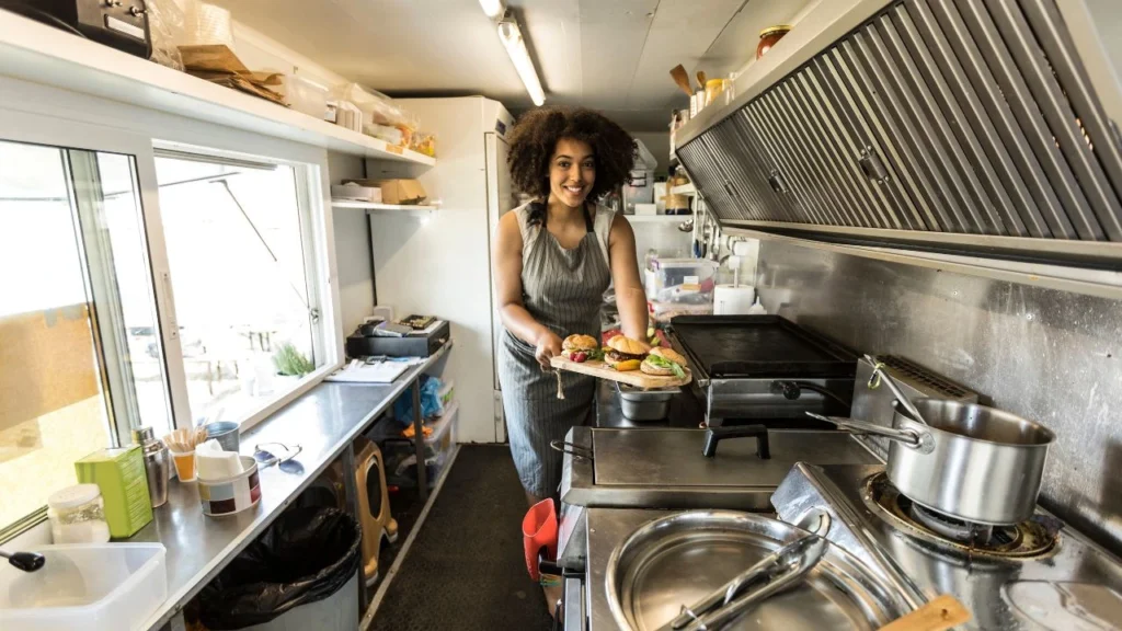 Smiling chef presenting burgers inside a food truck kitchen.